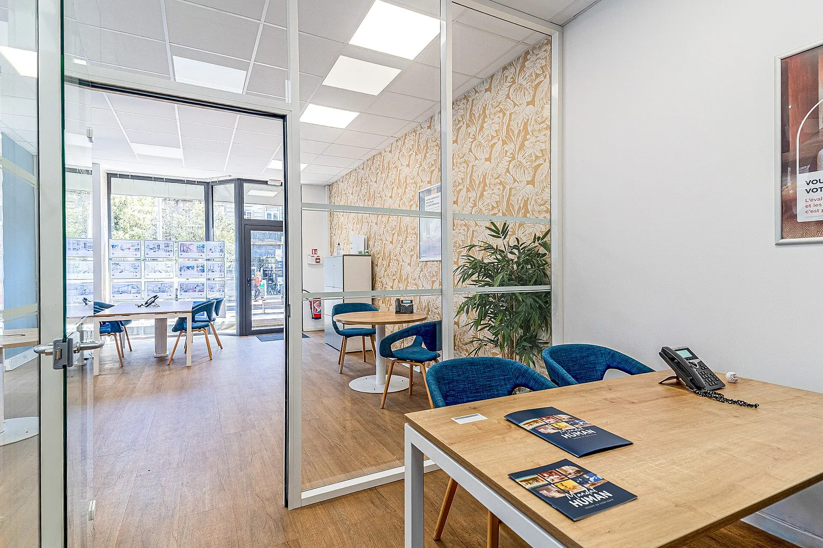 The bright inside of a French estate agency, with a glass partition, a wooden meeting table, blue chairs and listings pinned in the window