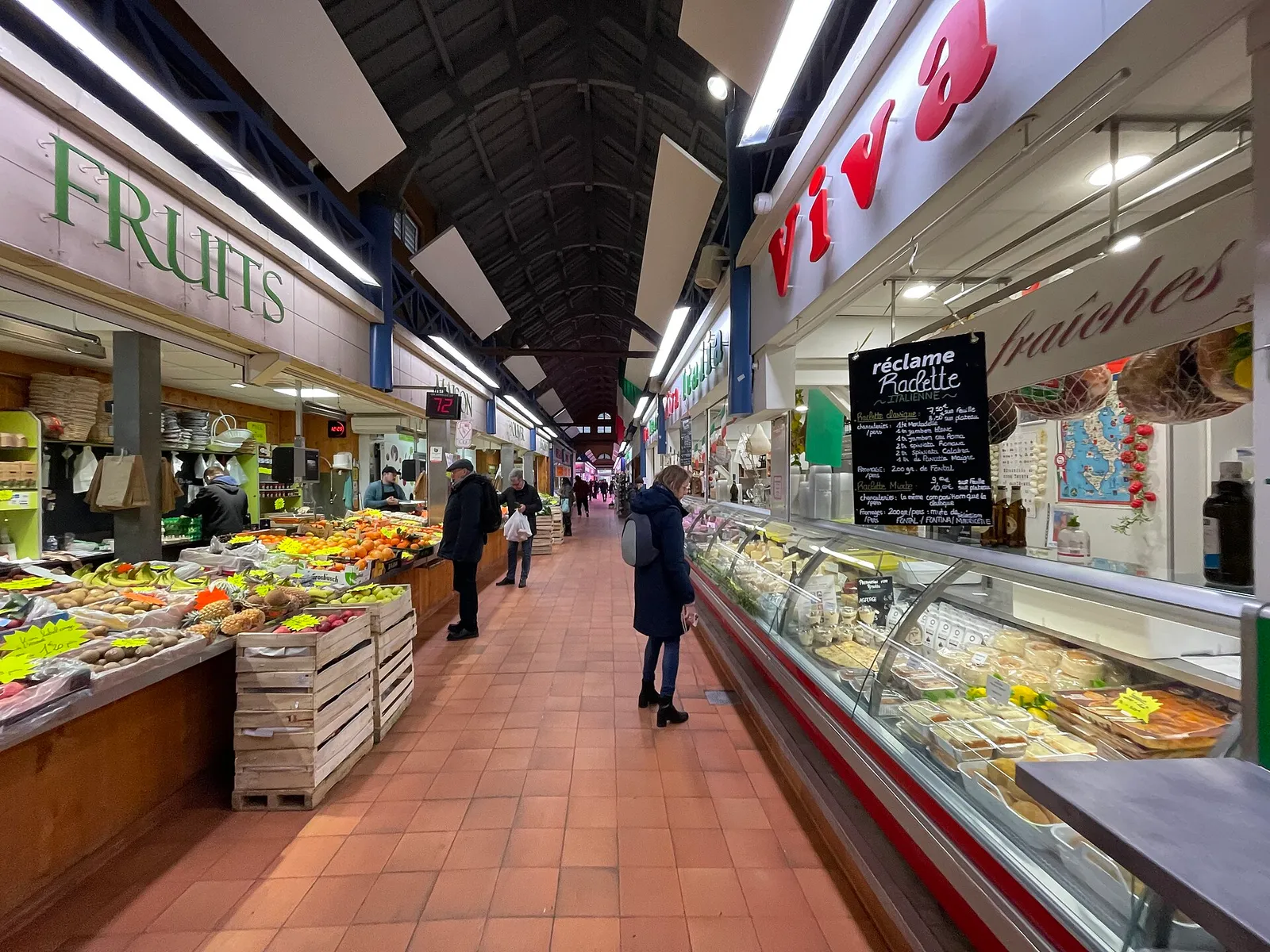 Shoppers in the covered market hall of Metz, between a fruit and vegetable stall and a delicatessen counter, on a red tile floor