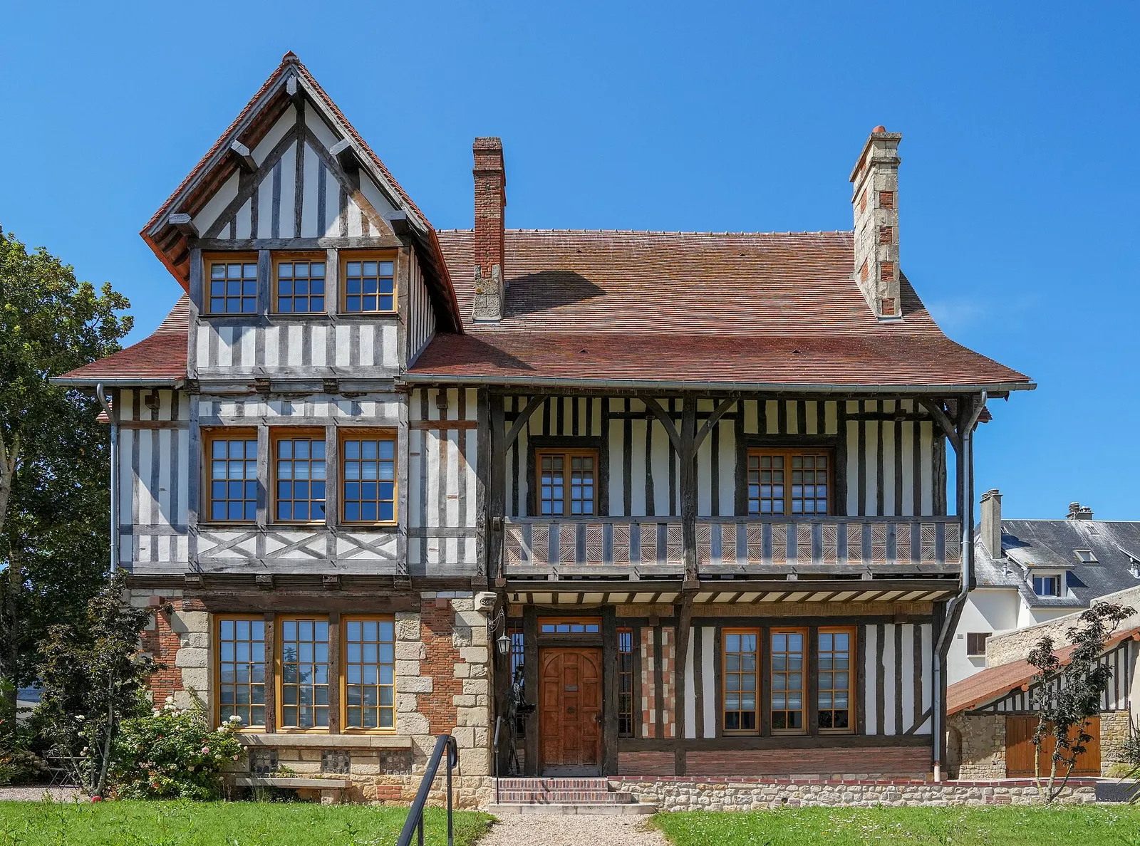 A half-timbered Norman villa in Deauville with a tiled roof, a wooden front door and a lawn in front, under a blue sky