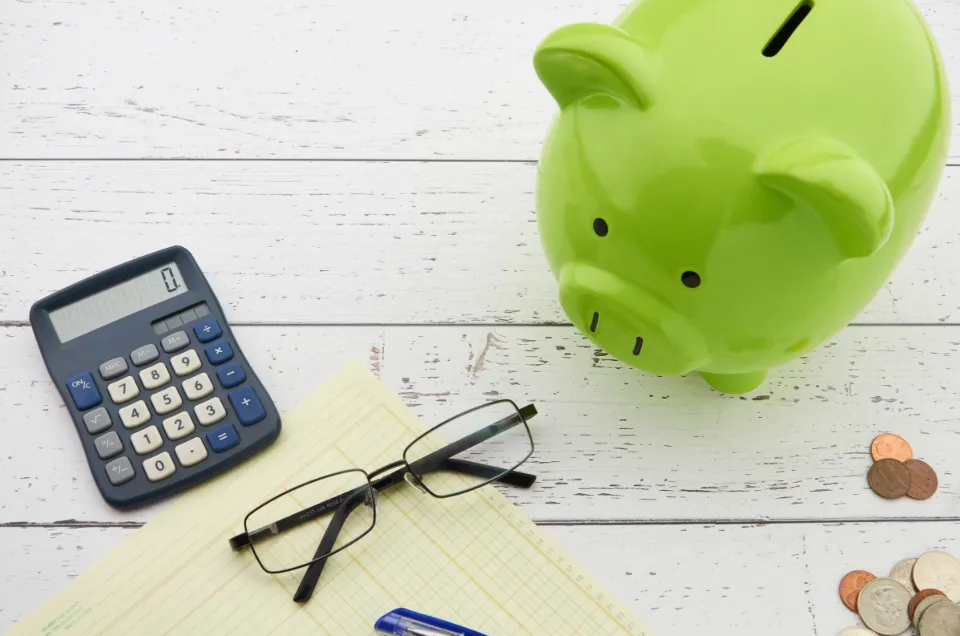 A green piggy bank beside a calculator, glasses and a few coins on a white wooden table
