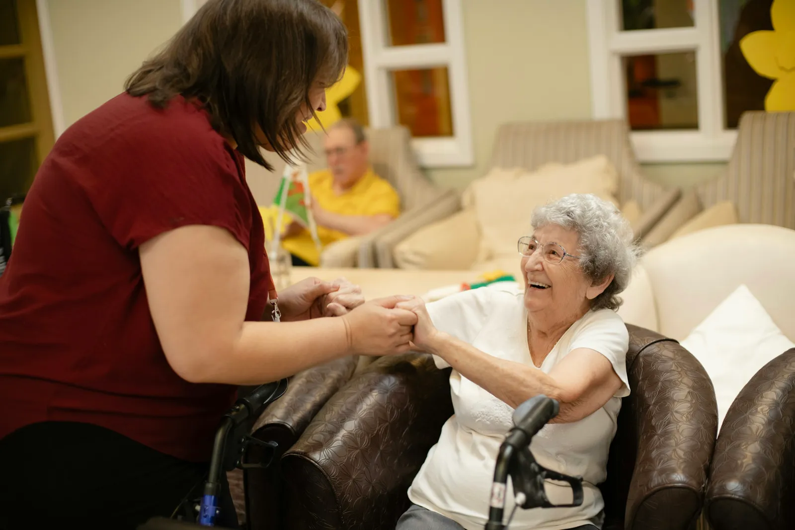 A care worker in a maroon shirt holds the hands of a smiling elderly woman seated in an armchair, with her walker in the foreground and a day room behind them