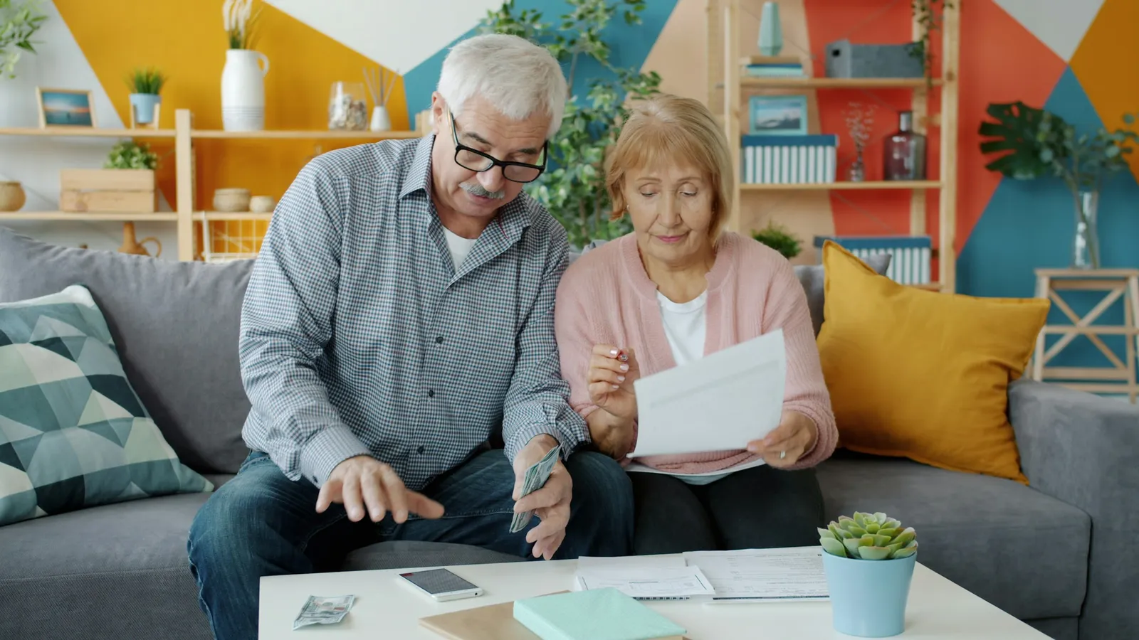 An older man in glasses and a woman in a pink cardigan sit on a grey sofa studying a letter and a few banknotes, with papers and a phone on the coffee table in front of them