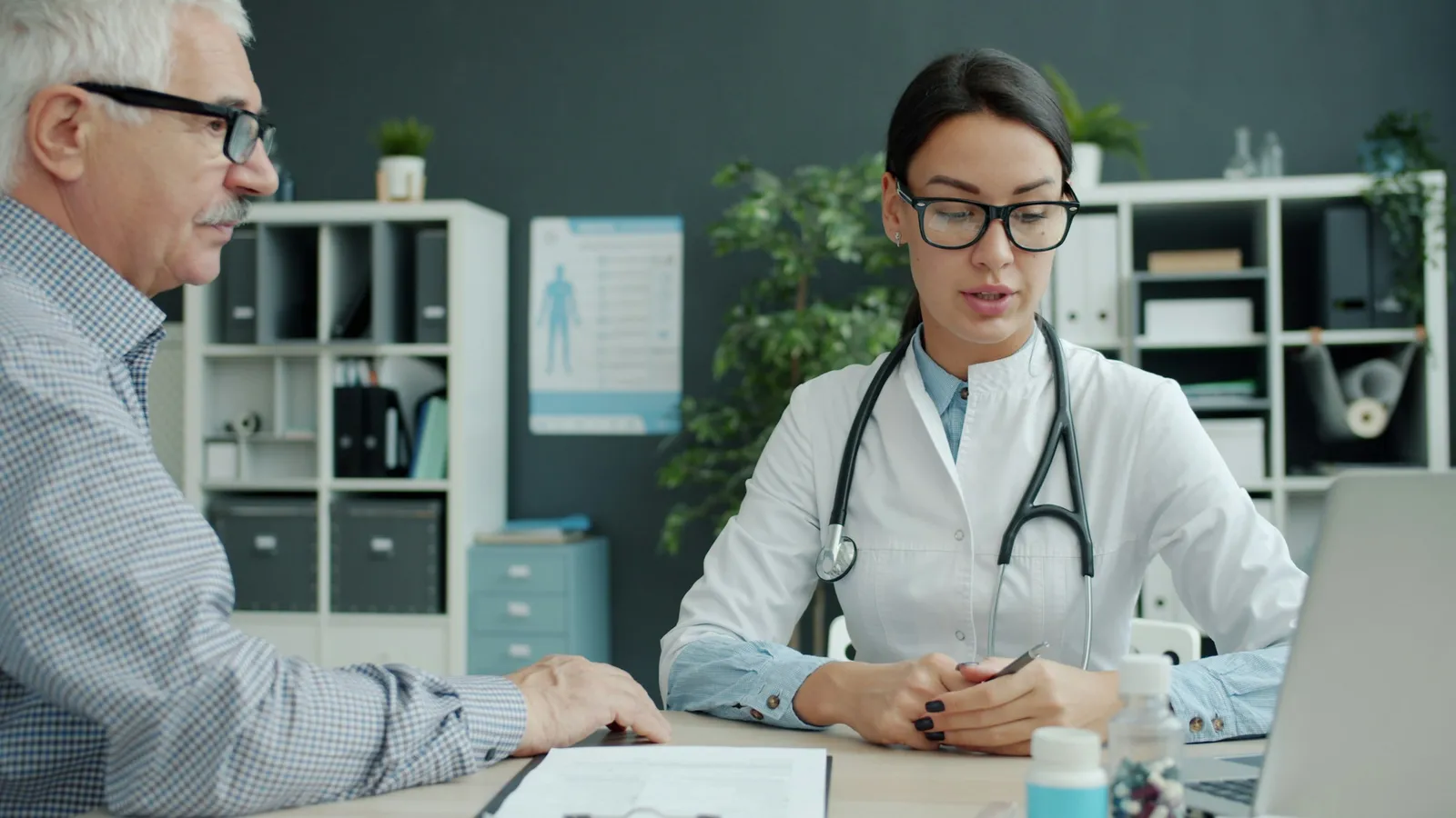 A woman doctor with a stethoscope and glasses speaks to an older man across a desk, with paperwork, a laptop and medicine bottles in front of them