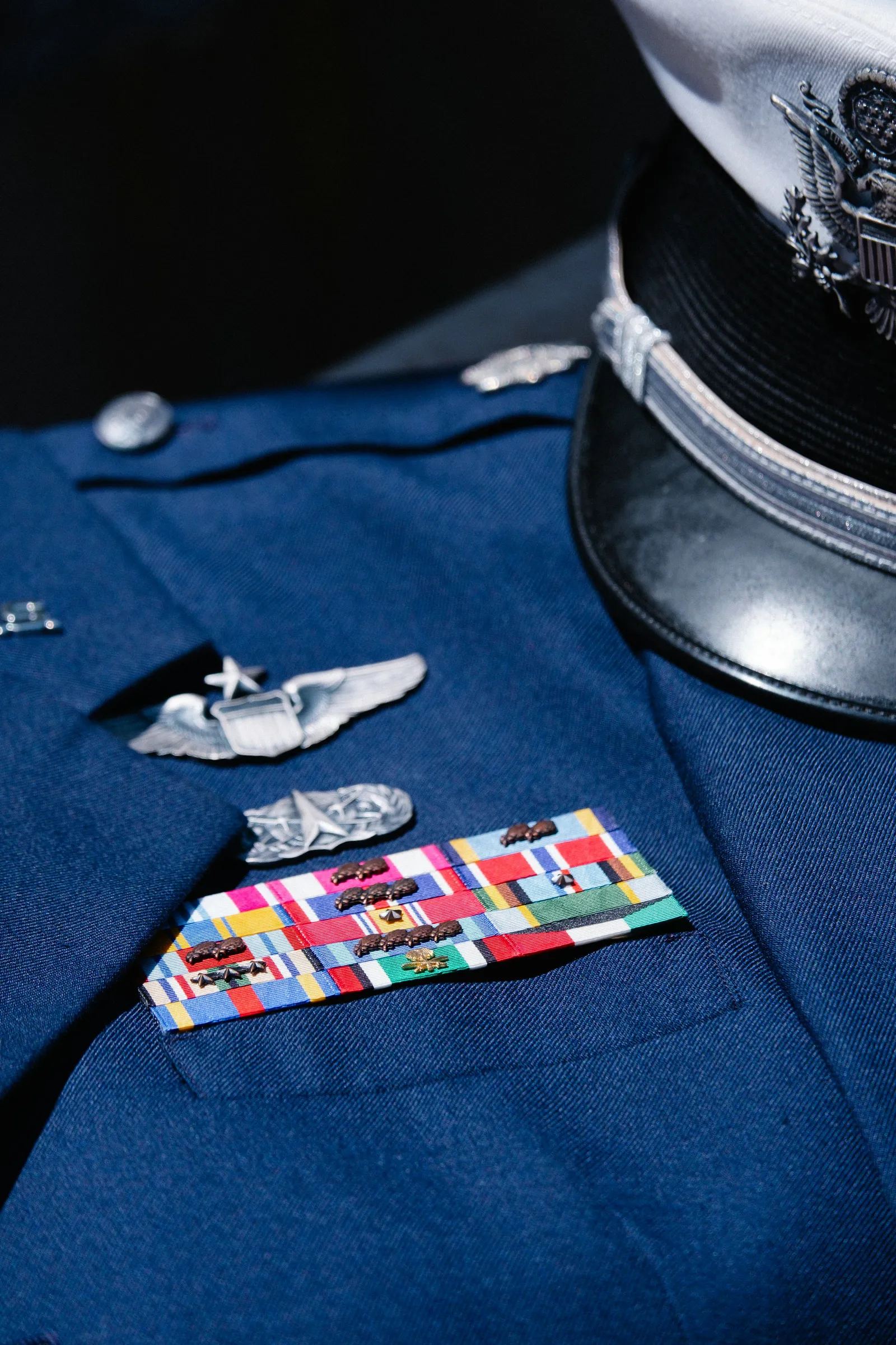 A blue military dress uniform jacket with silver pilot wings, rows of colourful service ribbons and a white officer's cap resting on the shoulder