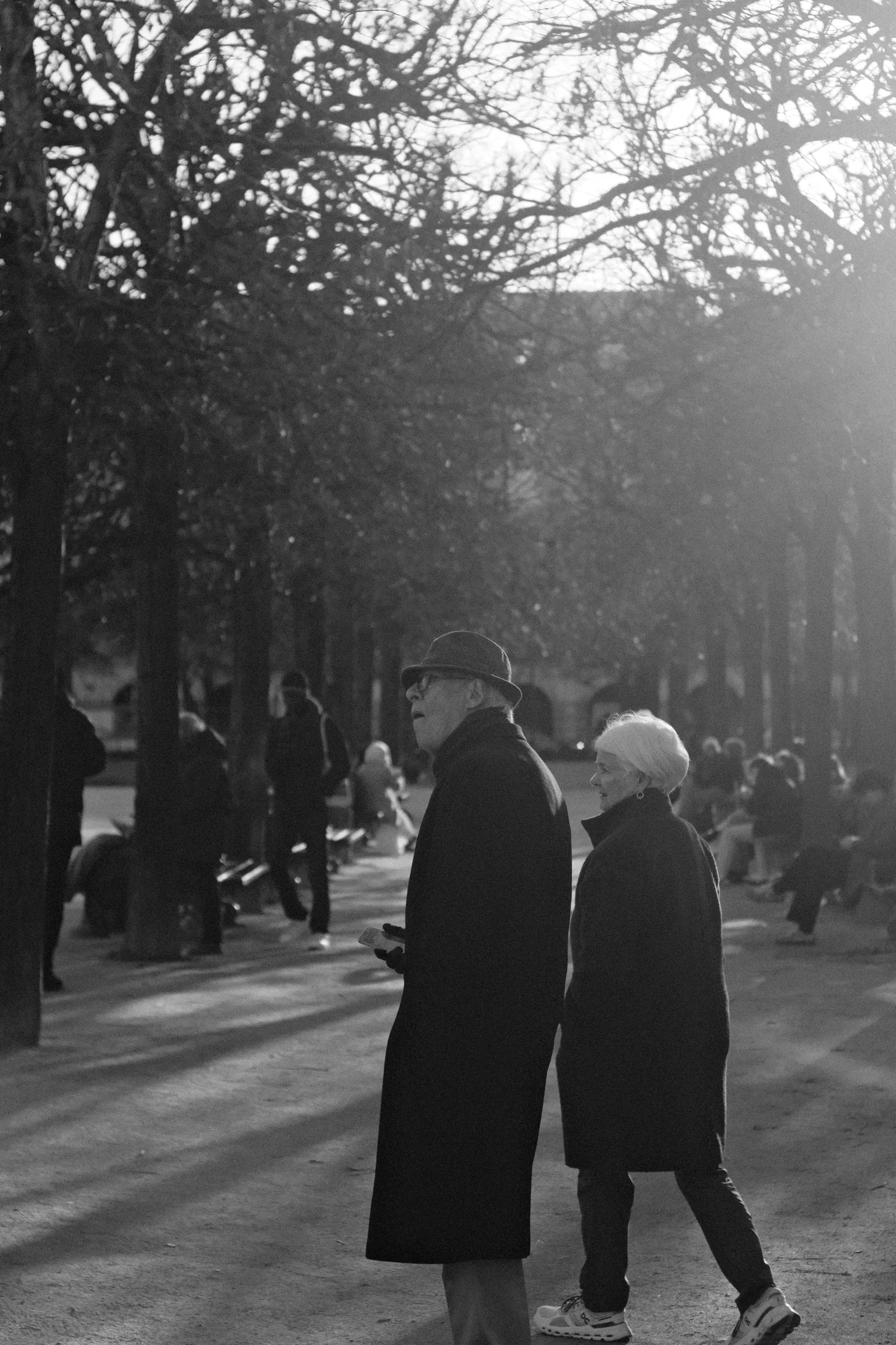 An older man in a hat and dark coat and a white-haired woman walk side by side along a tree-lined path in a park, photographed in black and white