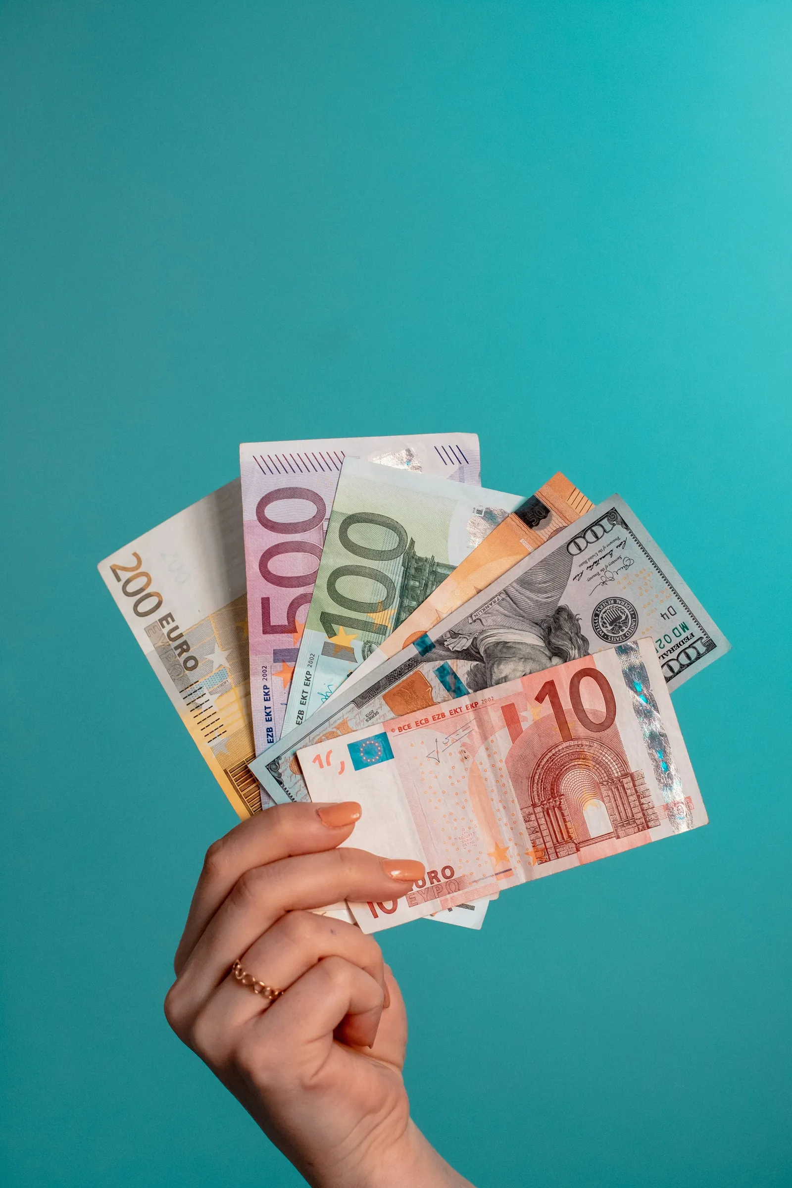 A hand holding a fan of euro and US dollar banknotes against a teal background
