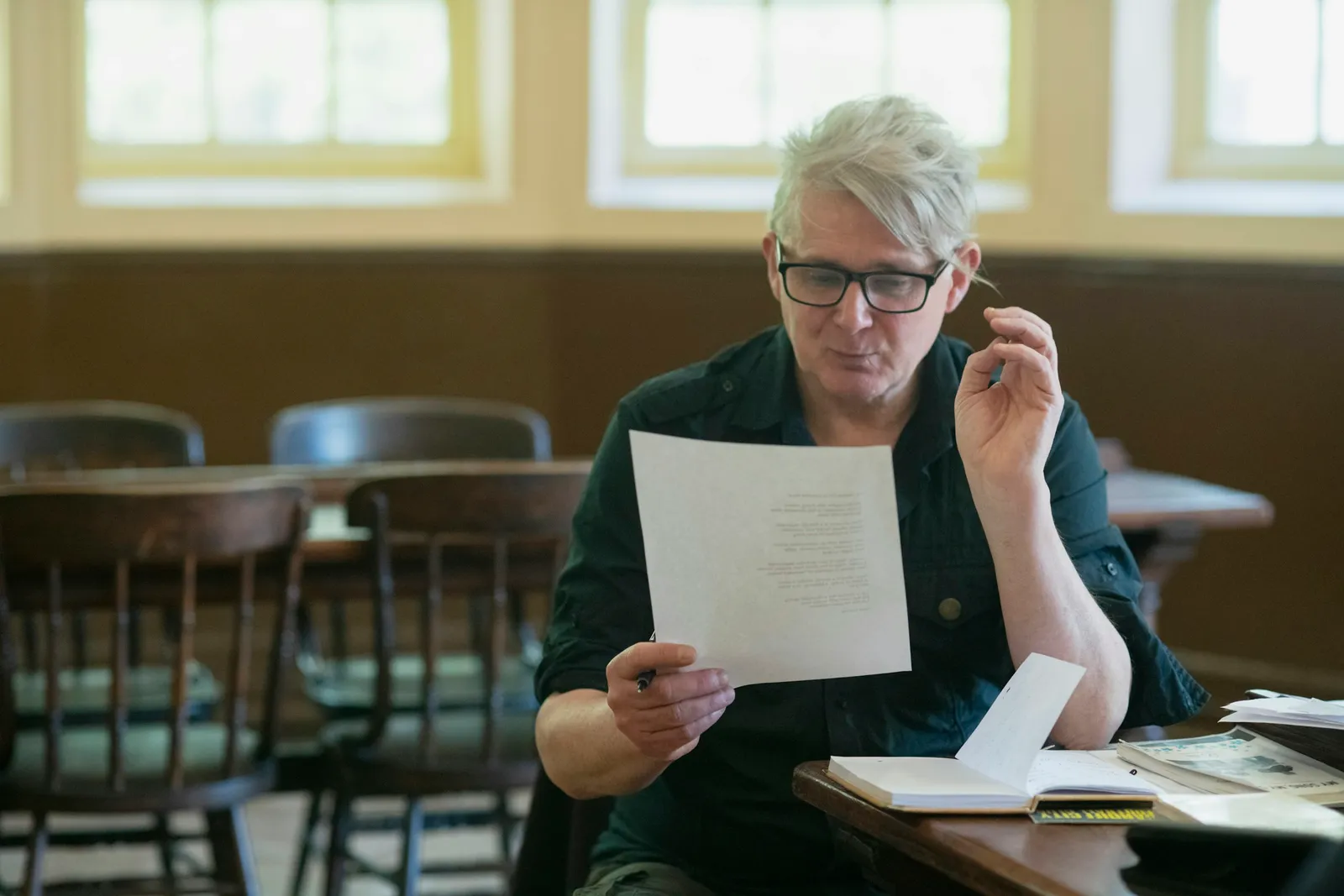A man with grey hair and dark-framed glasses sits at a wooden table in a bright room, reading a typed sheet of paper with a notebook open in front of him