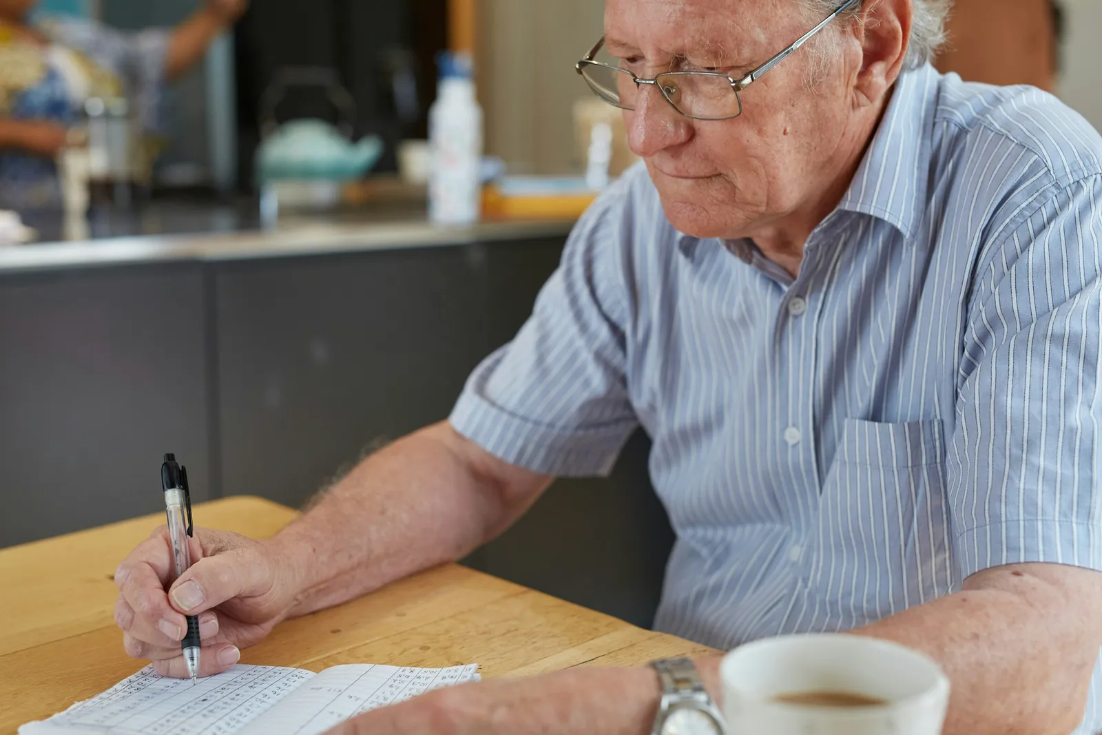 An older man in glasses and a striped shirt writes by hand on a sheet of paper at a wooden table, a cup of coffee in front of him