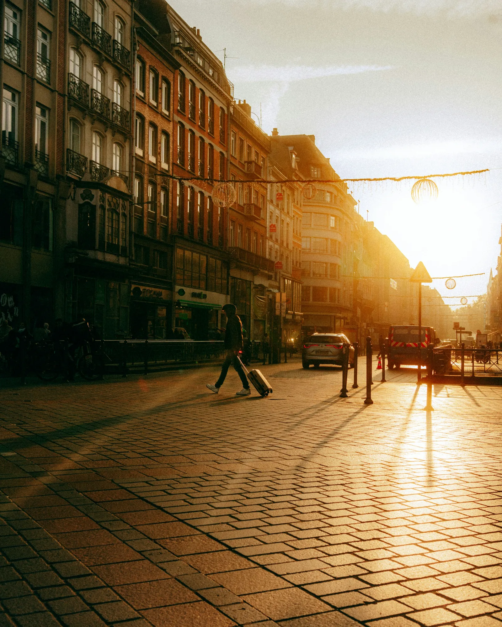 A traveller pulls a wheeled suitcase across a sunlit paved street between old buildings
