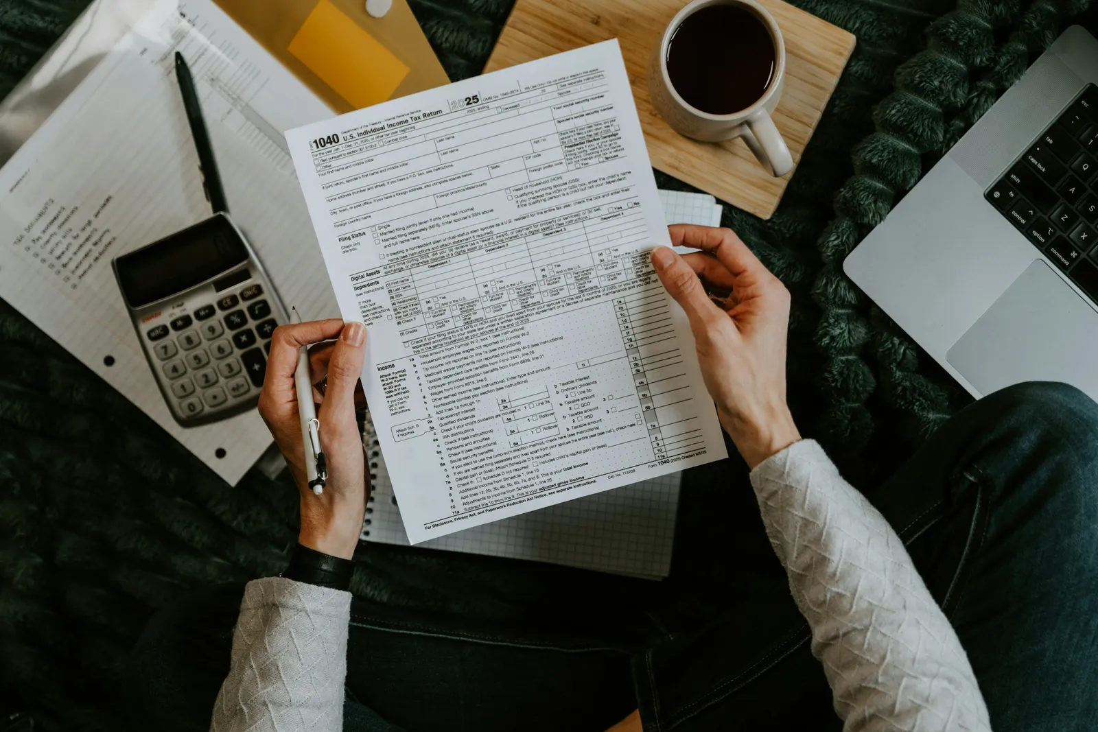 A person holding a printed US Form 1040 above a desk with a calculator, a pen, a notebook and a laptop